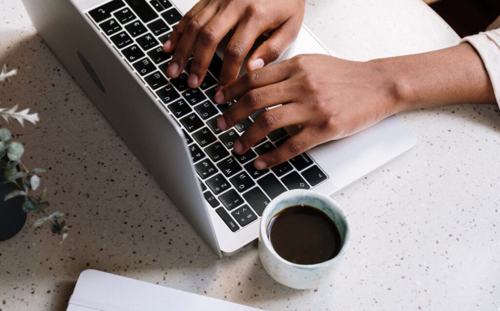Hände einer Person, die auf der Tastatur eines Laptops tippen, mit einer Tasse Kaffee daneben.